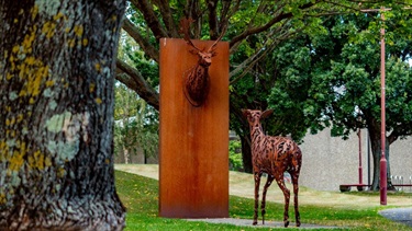 A doe looks up at the head of a buck mounted on a steel block set amongst trees in a small park.