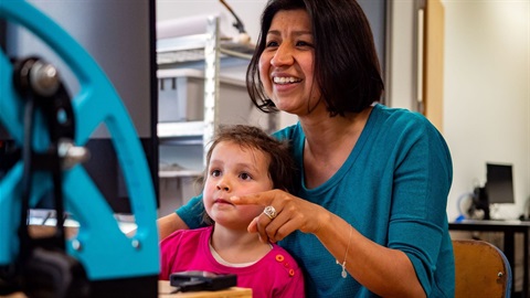 Smiling woman with child on her lap sitting at one of the makerspace machines.