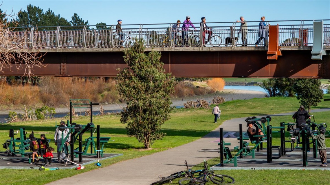 People working out on the gym equipment lining the river pathway while cyclists cross the bridge above them.