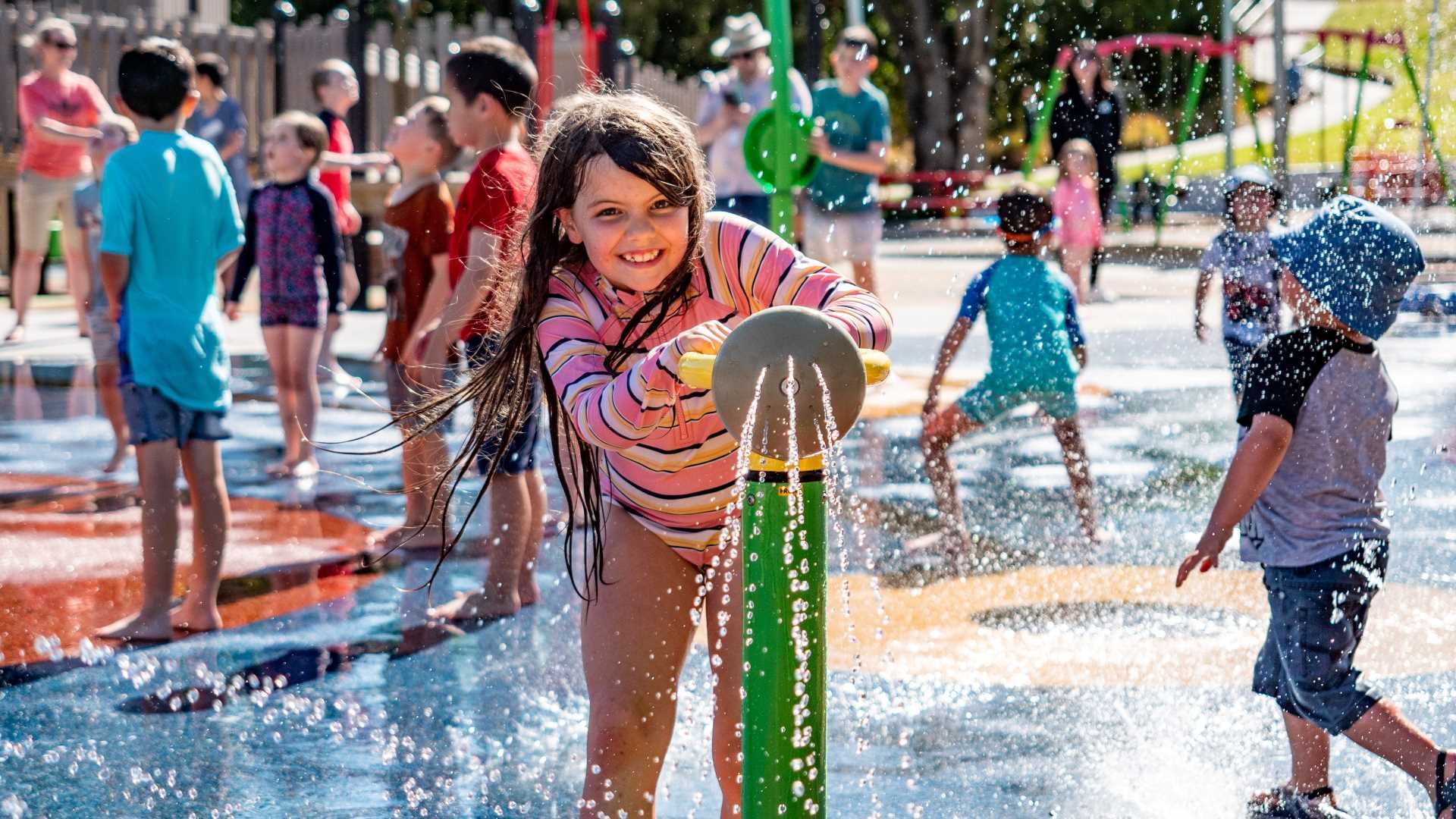 A girl in togs grins in the water as other children splash around her.