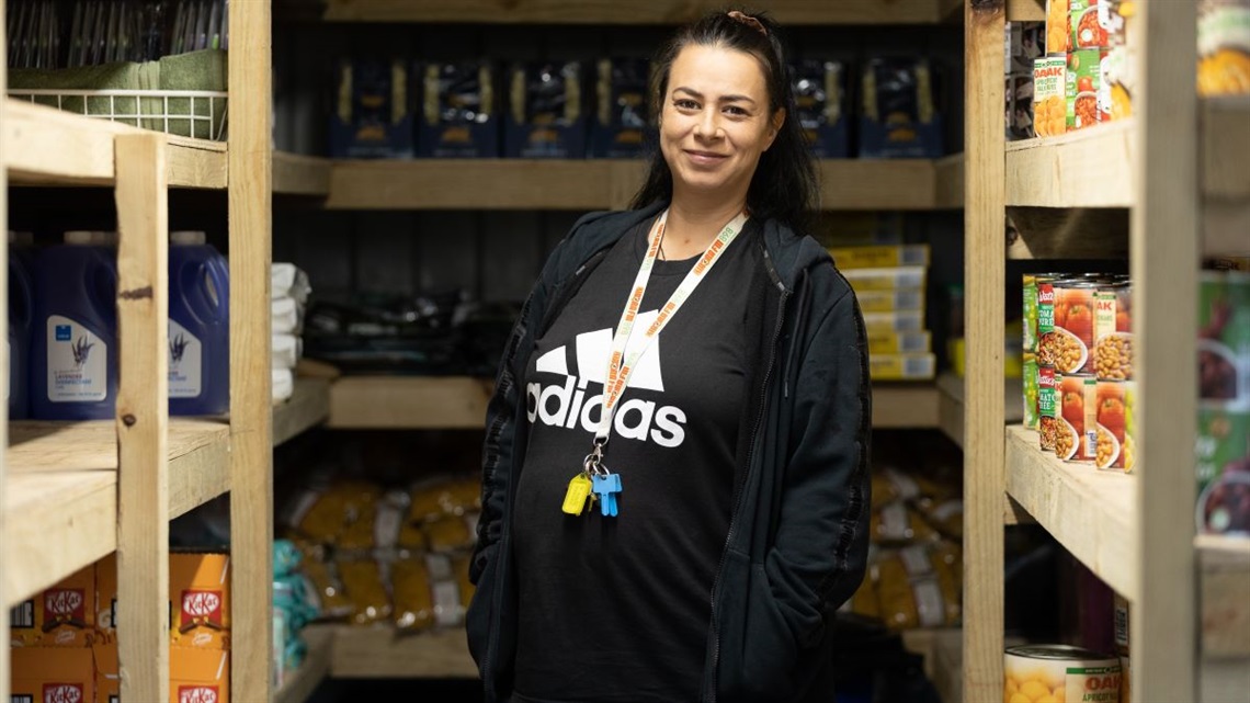 A smiling woman standing inside a community pantry with well-stocked shelves.