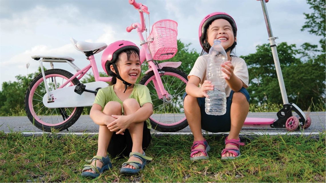 Two tamariki grin ear to ear as they pause from scooting and biking along the river pathway. One is drinking from a large water bottle.