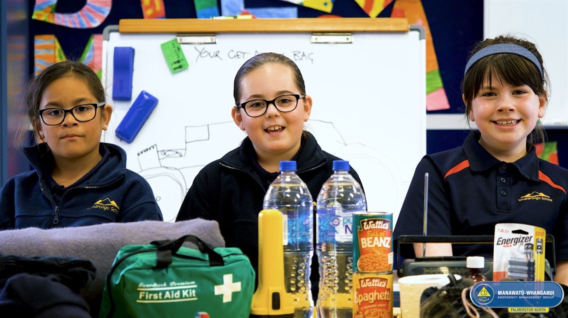 Three tamariki preparing an emergency getaway bag with food, batteries, water, first aid kit, batteries, a radio and a blanket.