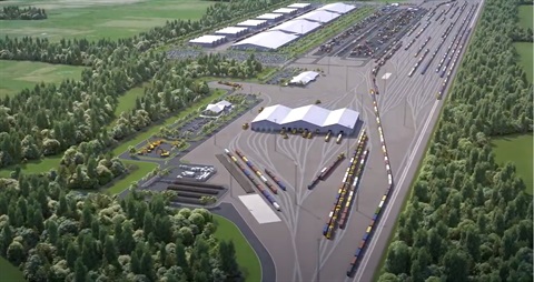 Photo shows huge concrete area with tracks, trains, trucks and sheds, surrounded by green space and trees.