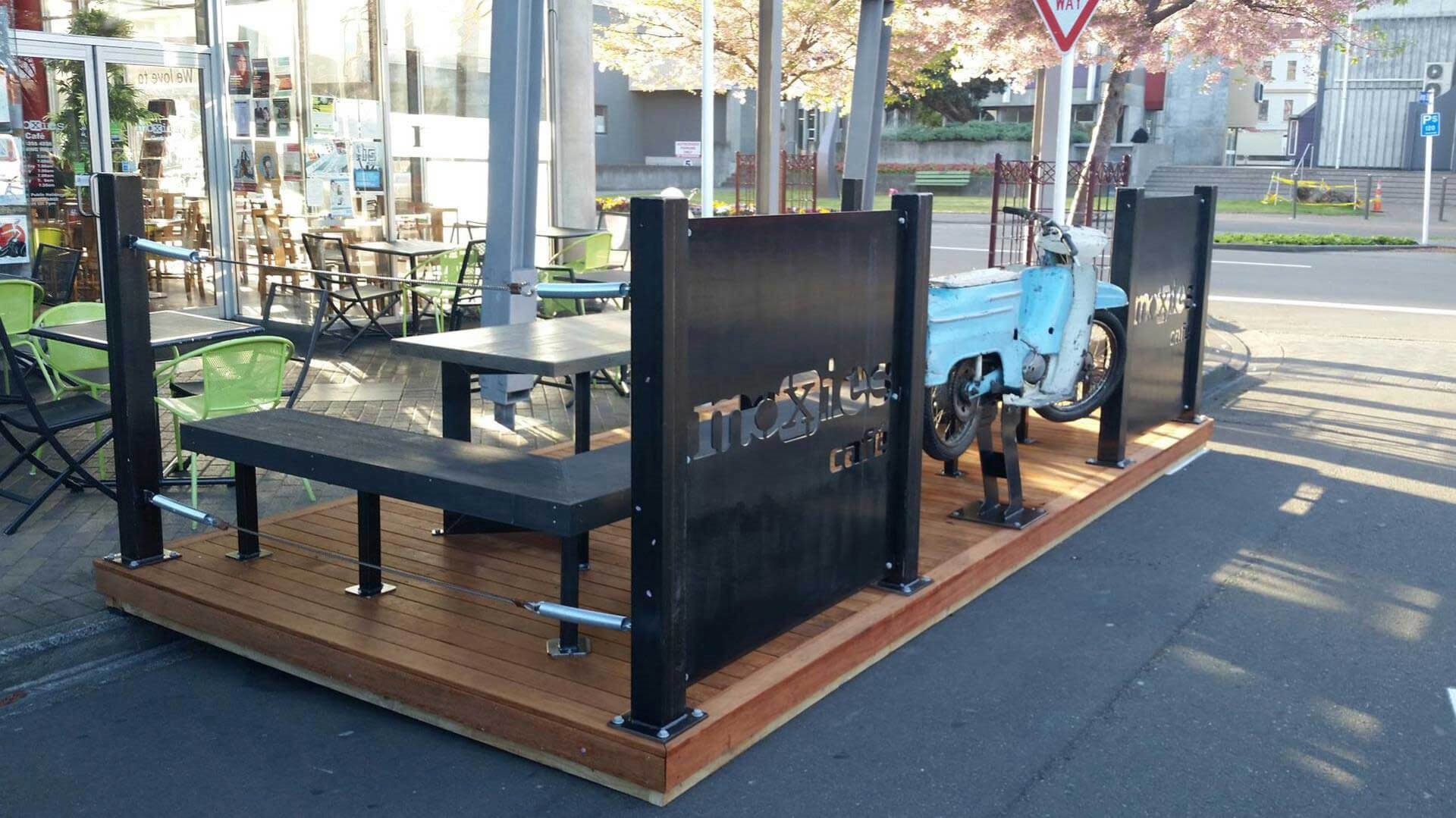 Carpark that has been converted into a shared social space, with chairs, tables, and a motorbike sculpture.