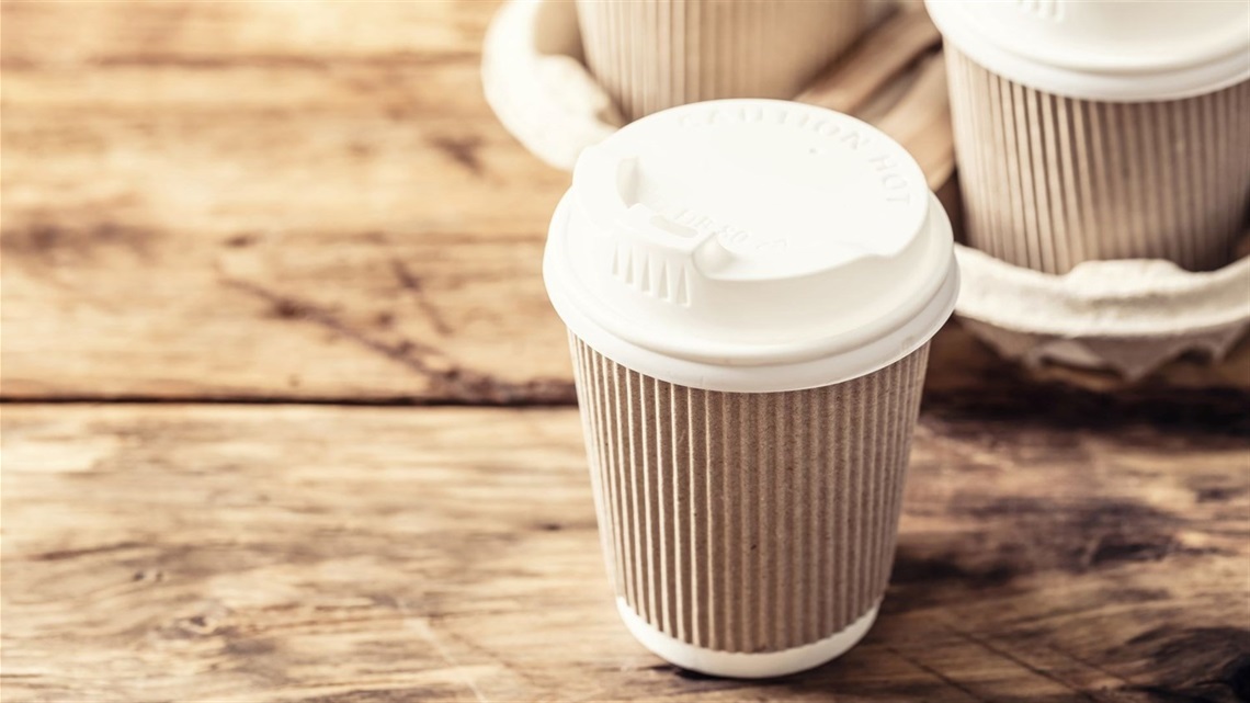 Takeaway coffee cups with plastic lids in a cardboard carrier on a wooden tabletop.