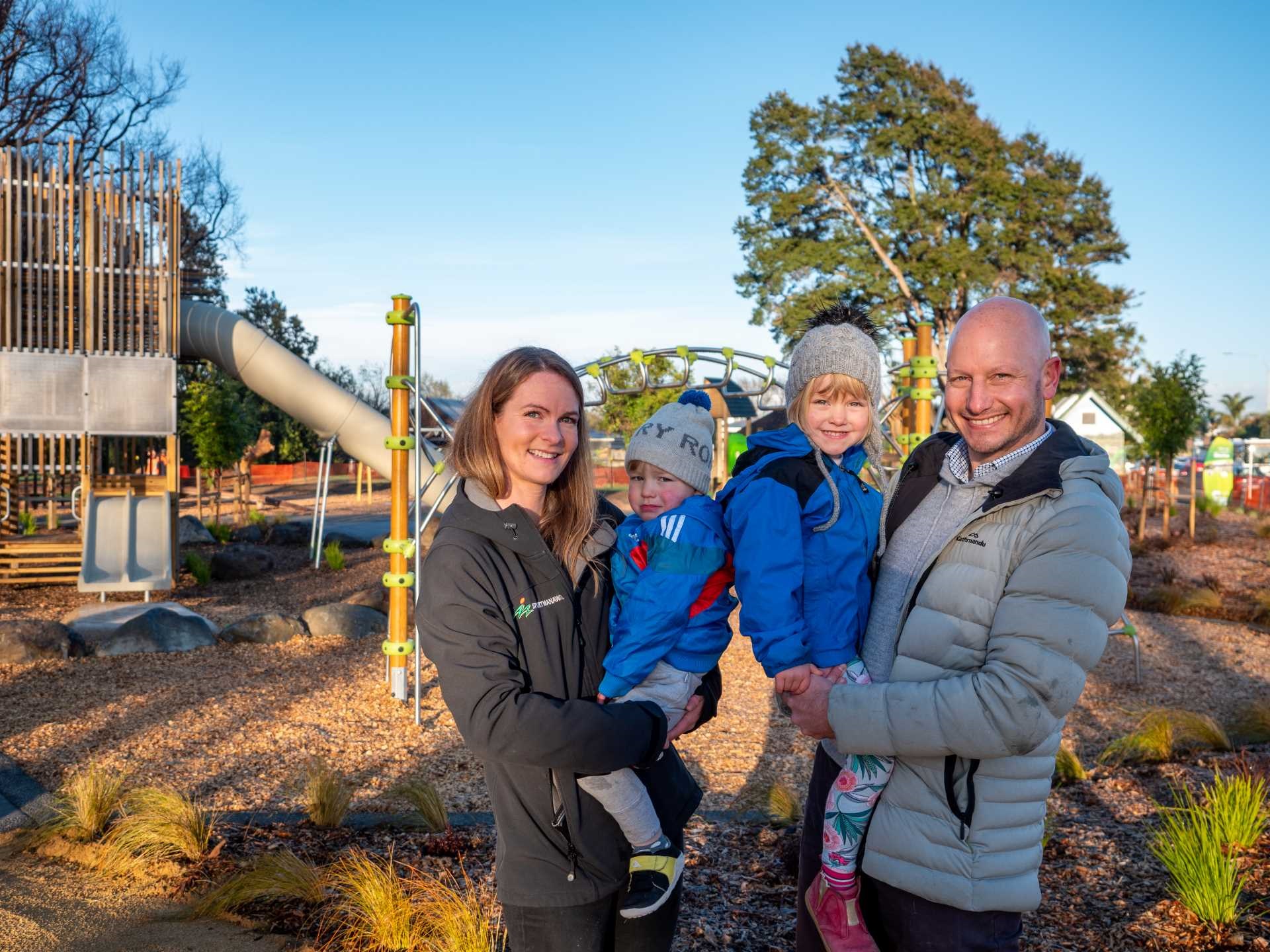 Photo shows couple with their two young children and a playground in the background featuring a fort and slide.